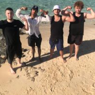 Vier junge Männer halten ihre Muskeln in Höhe und stehen barfuß am Strand