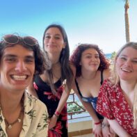 vier Personen lächeln strahlend auf einem Balkon, im Hintergund ist das Meer, blauer Himmel und Palmen zu sehen.